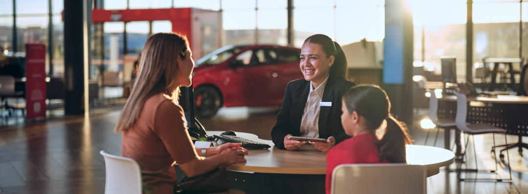 A woman in a blazer with a namebadge sits at a round table smiling at a woman and a young girl inside a Toyota dealership.