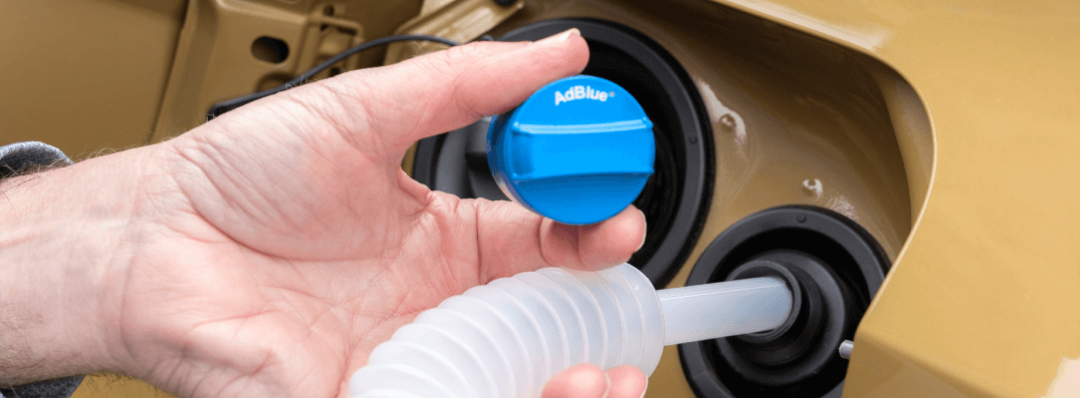 A hand holding a blue "AdBlue" cap while a clear plastic nozzle is inserted into the vehicle's fluid filler port.