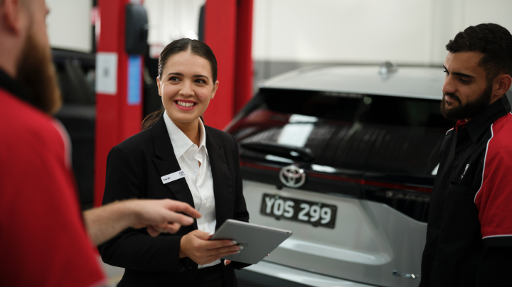 A woman holding a tablet and smiling while talking to two men in a workshop next to a silver Toyota.