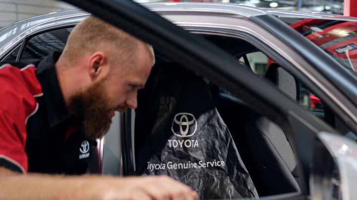 A man in a red and black Toyota uniform looks inside a silver vehicle with a Toyota Genuine Service protective cover placed on the driver's seat.