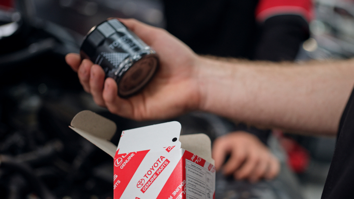 A hand holds a black cylindrical car part above an open red and white Toyota box.