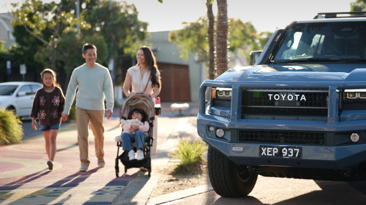 A blue Toyota Prado is parked on a suburban street. A man, a woman, a young girl, and a baby in a pram walk on the footpath in the background.