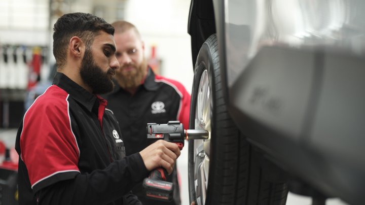 A man in a red and black Toyota uniform uses a rattle gun to secure a wheel on a hoisted car. Another man looks on in the background.