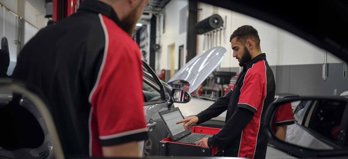 A man in a red and black Toyota uniform uses a laptop on a red cart next to a car inside a workshop. Another man stands in the foreground.