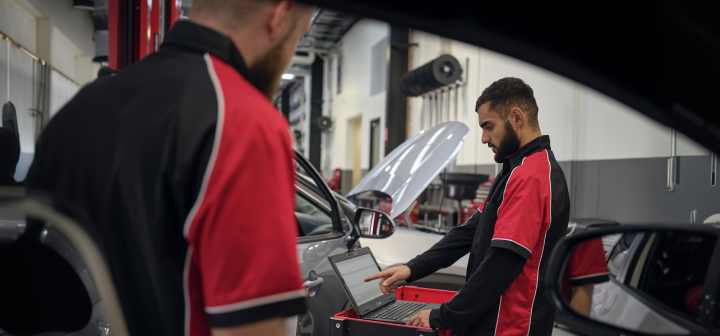 A man in a red and black Toyota uniform uses a laptop on a red cart next to a car inside a workshop. Another man stands in the foreground.