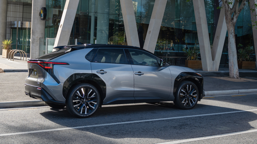 A silver Toyota bZ4X is parked on the side of a city street in front of a modern building with large concrete pillars.