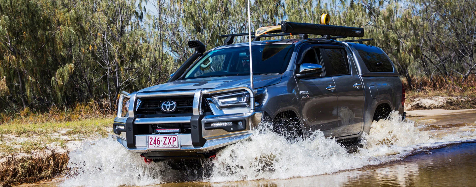 A grey HiLux fitted with a Toyota Genuine Bull Bar, driving through a shallow river in the Australian bush.