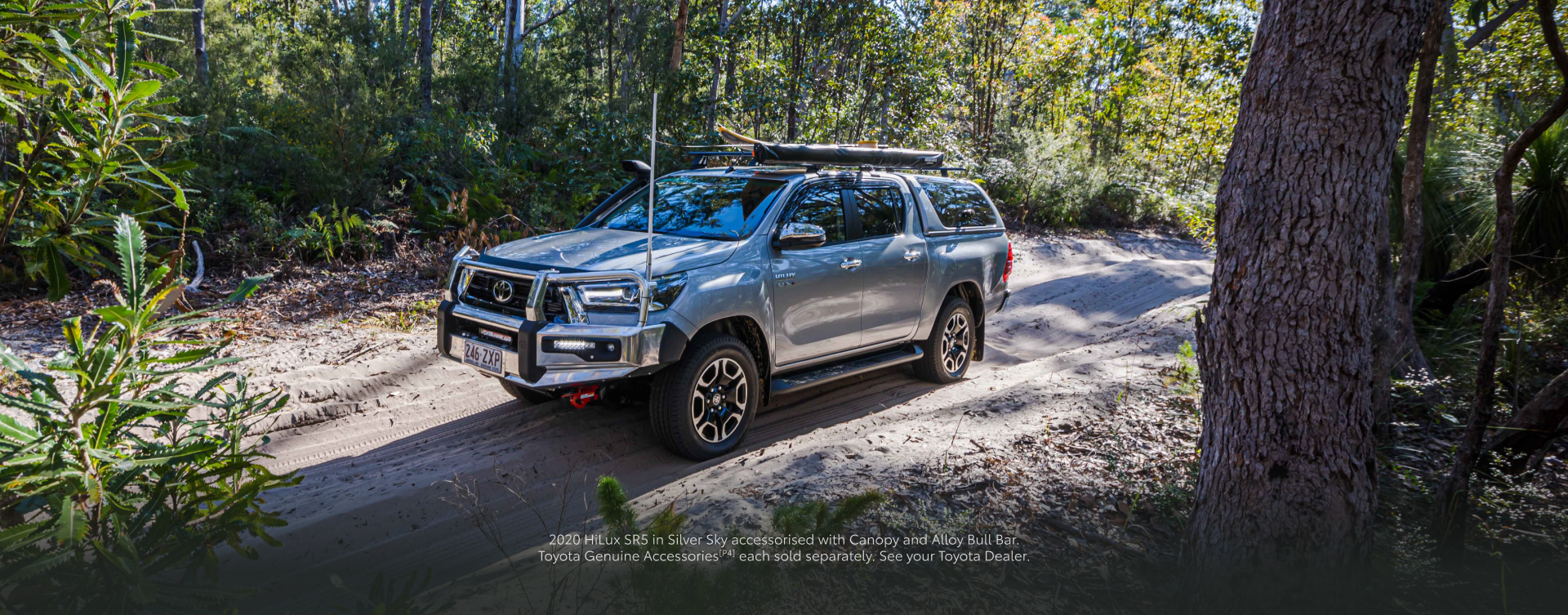 A Toyota HiLux in Silver Sky, driving along a dirt road in a forested coastal area. The vehicle is equipped with a Toyota Genuine Roof Rack, to which surfing and fishing gear are attached.