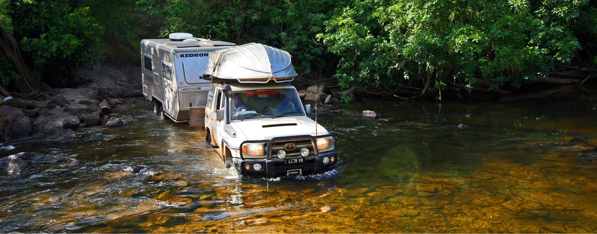 A white Toyota LandCruiser driving through a river crossing, a caravan in tow. A small boat is mounted on the roof rails.