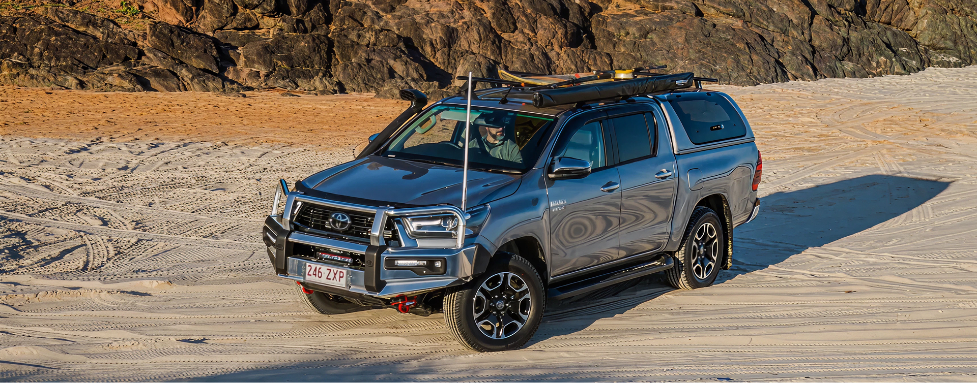 A Toyota HiLux in Silver Sky, parked in front of a rocky embankment on a sandy beach at sunset. It’s equipped with a Toyota Genuine Roof Rack, with surfing and fishing gear mounted.