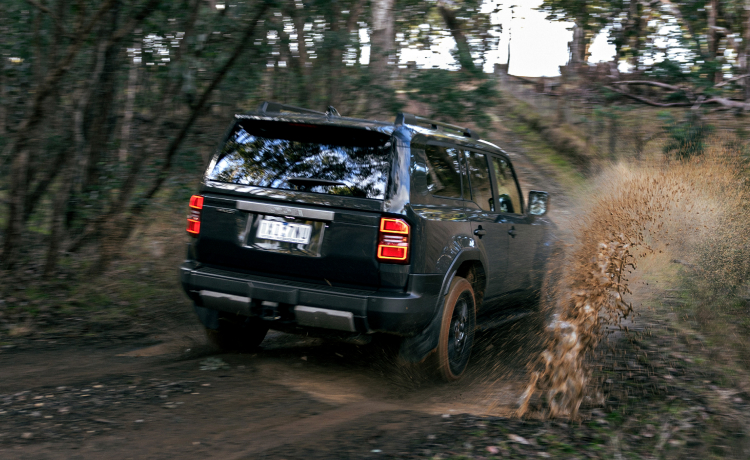 A Toyota LandCruiser Prado in Eclipse Black, driving down a dirt road in the bush, kicking up mud and water.
