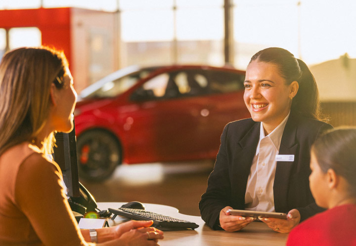A woman in a showroom focuses on a digital tablet while a consultant points to the screen, discussing vehicle details or financing options.