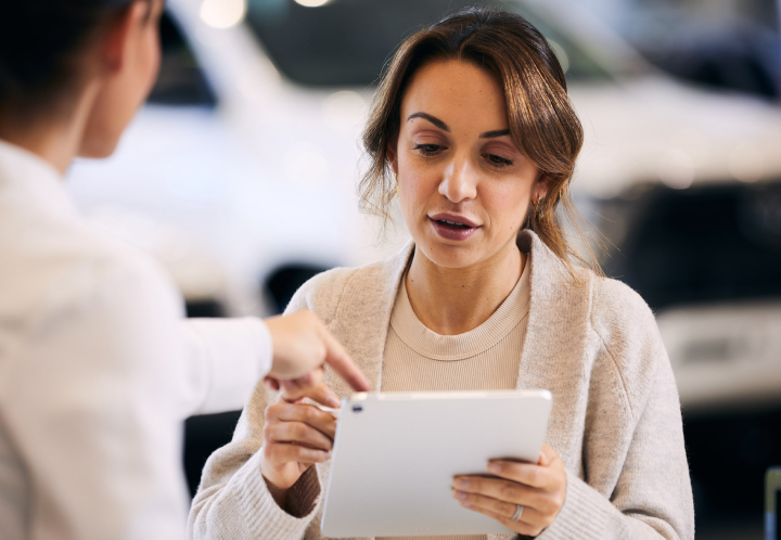A woman in a showroom focuses on a digital tablet while a consultant points to the screen, discussing vehicle details or financing options.