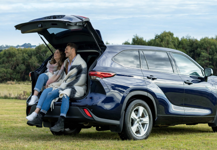 A smiling couple wrapped in blankets sits in the open cargo area of a dark blue SUV, enjoying an outdoor view in a rural meadow setting.