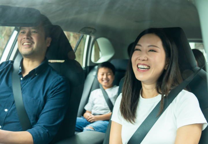A cheerful family of three enjoys a road trip inside a modern vehicle, with the mother laughing in the foreground and the father and son visible in the background.