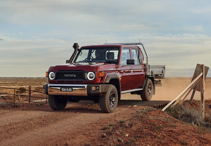 A red Toyota Land Cruiser 70 Series flatbed truck drives across a dusty, unpaved road in a vast, open outback environment.