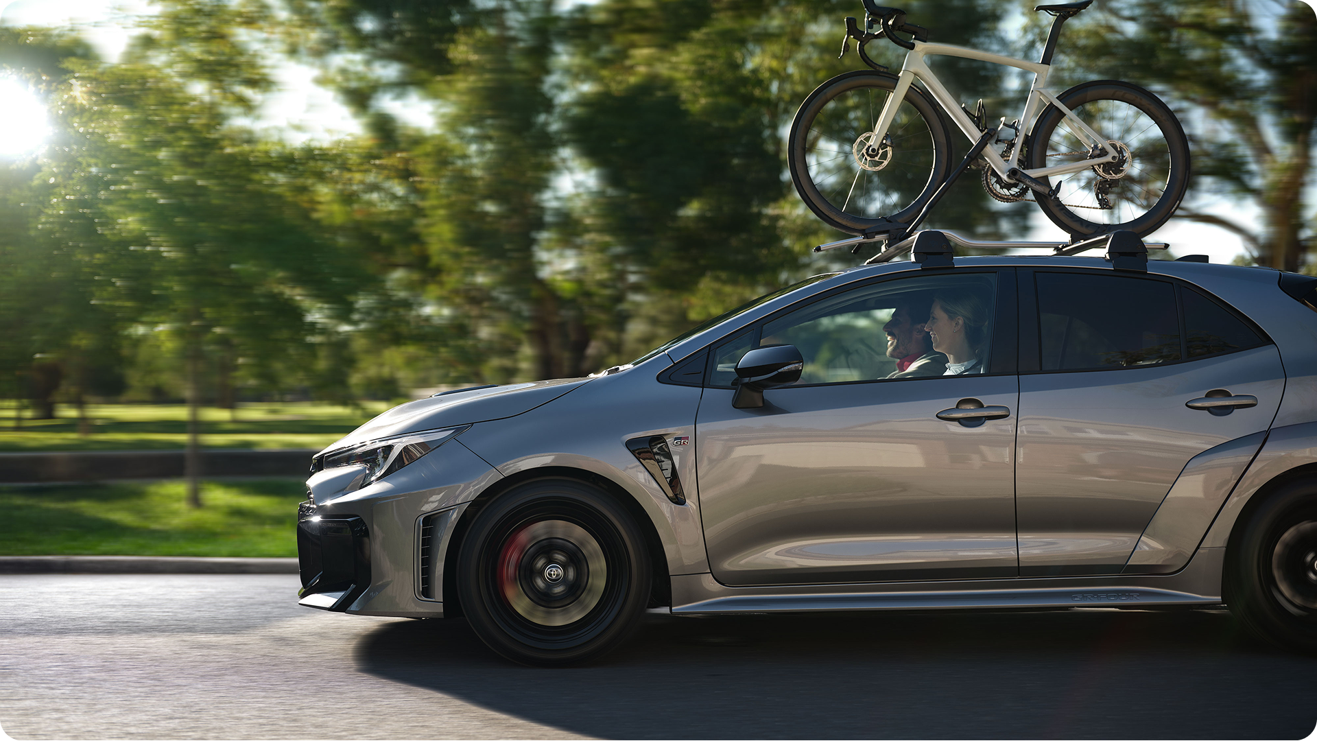 Profile view of a silver Toyota GR Corolla hatchback driving past green trees, equipped with a roof rack holding a road bicycle.